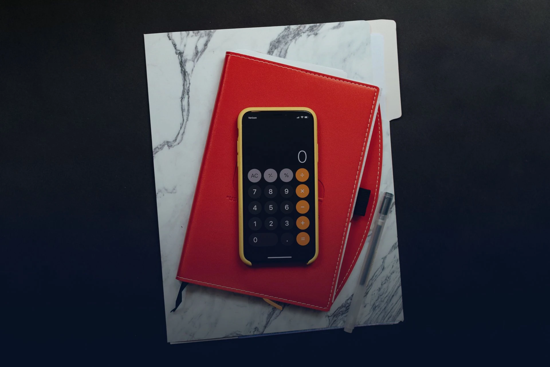 Iphone appoggiato sopra ad una agenda rossa che a sua volta è appoggiata su un computer con cover con disegno di marmo