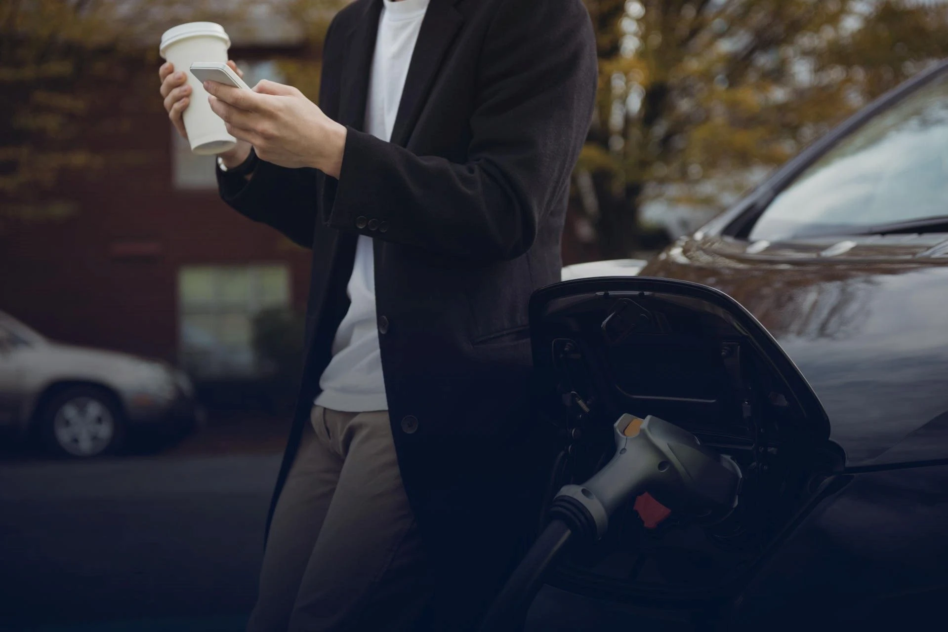 Ragazzo con in mano caffè e smartphone appoggiato ad un'auto plug-in che è in carica
